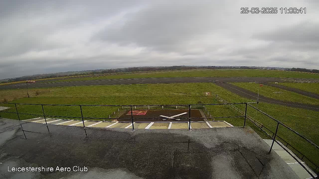 A view from a webcam stationed at the Leicestershire Aero Club shows a wide, open landscape under a cloudy sky. In the foreground, there is a railing surrounding a viewing platform, and the ground is wet, indicating recent rain. Below, a small aircraft painted in white is positioned on the tarmac, with various patches of grass and a few runways visible in the background. The overall ambiance is calm and quiet, typical of an airport environment.