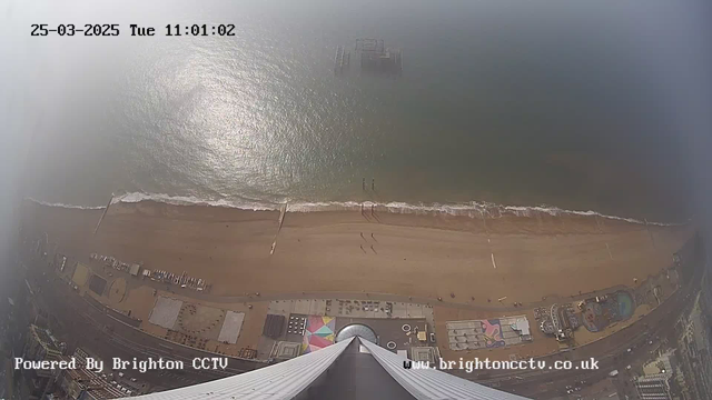Aerial view of a sandy beach lined with a few colorful beach huts. The waves gently lap at the shoreline, and a pier extends into the sea. There are minimal people on the beach, and a distant amusement park can be seen to the right, featuring rides and a pool. The sky appears slightly overcast, with a foggy atmosphere. The date and time are displayed in the top left corner.
