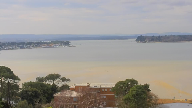 A scenic view of a calm body of water under a mostly cloudy sky. The foreground features some greenery with trees, and a low brick building with multiple windows. In the background, a shoreline is visible, dotted with boats and various structures. The landscape includes gentle hills and a distant tree line that defines the horizon. The colors are soft, with a mix of muted greens, browns, and blues, suggesting a tranquil atmosphere.
