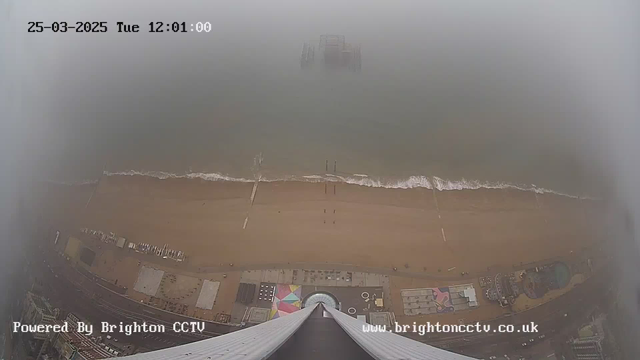 An aerial view of a beach scene taken from a high vantage point. The image shows a sandy beach with gentle waves lapping at the shore. There is a pier partially visible in the foggy distance. The beach area includes various structures and possibly amusement rides, though details are obscured by fog. The sky appears grey, indicating overcast weather conditions. The lower part of the image shows sections of colorful designs, likely from beachside attractions or mats.