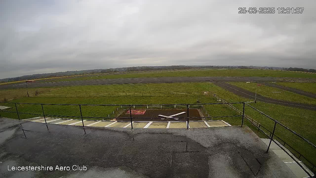 A view from a high vantage point overlooking an airfield. The foreground features a flat rooftop with a railing, and below, there is an airstrip bordered by green grass. In the center of the image, there is a small model airplane outlined on the ground. The sky above is cloudy, and the overall atmosphere is calm. The time display shows 12:01 on March 25, 2025, and the name "Leicestershire Aero Club" is visible at the bottom.