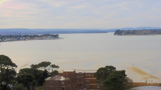 A panoramic view of a calm body of water, with light reflections making the surface shimmer. The shoreline features a small harbor with several boats and a few buildings visible on the left side. In the foreground, there are trees and a building, while a lone figure stands on a sandbar in the distance. The background shows rolling hills and a cloudy sky, creating a tranquil atmosphere.
