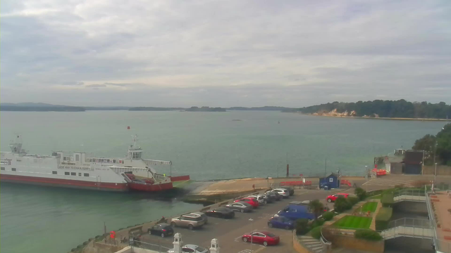 A ferry is docked at a harbor with a light greenish-blue body of water. Numerous vehicles, including red, silver, and dark blue cars, are parked along the waterfront. The background features a coastline with trees, cliffs, and a distant shoreline. The sky is overcast with gray clouds.