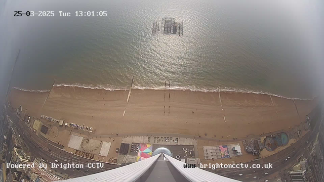 A high-angle view of a beach and shoreline, taken from a webcam. The sandy beach stretches horizontally across the image with small waves lapping at the shore. Several people are visible walking along the sand. In the foreground, various structures including colorful beach huts and a circular pattern on the ground can be seen. A pier extends into the water, and a section of the ocean is shimmering in the sunlight. The date and time are displayed in the top left corner.