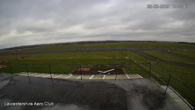 A cloudy sky covers the scene with soft gray tones. Below, a flat expanse of grass stretches across the landscape, bordered by a runway in the background. The runway has a haphazard arrangement of light and dark asphalt patches, with some markings visible. In the foreground, a railing is visible, suggesting the image is taken from a height, possibly from a rooftop or lookout point at an aero club. A small, white structure resembling an airplane is parked on the ground amidst the grass area. The overall atmosphere appears calm and open, with no visible movement.
