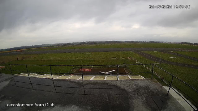 A cloudy sky hangs over a large expanse of green grass and a runway. In the foreground, there is a flat rooftop area with a railing. A small white aircraft is positioned on a marked area on the ground. The runway extends into the distance, bordered by a wooden fence. The atmosphere appears calm and still.