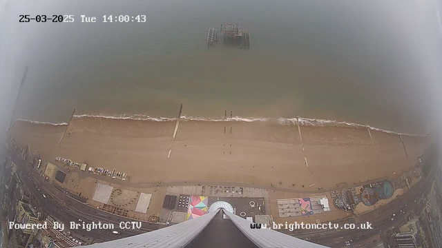 A high-angle view of a sandy beach with the sea visible in the background. The shoreline features gentle waves lapping against the sand. There is a pier extending into the water on the left side of the image, with remnants of an old structure partially submerged. The area immediately in front of the camera shows a winding road lined with buildings, some of which have colorful designs. The image is somewhat foggy, reducing visibility. The bottom of the image includes a timestamp and website information, indicating it is from Brighton's CCTV.