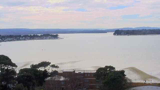 A view of a calm body of water extending to the horizon, with gentle waves creating a rippling effect on the surface. In the distance, low hills and trees line the shoreline, while a few buildings can be seen at the water’s edge. The sky above is a mix of soft pastel colors, indicating a cloudy day, with hints of blue peeking through. A small boat is visible on the water, along with a few floating markers. The foreground features some greenery and rooftops of nearby buildings.