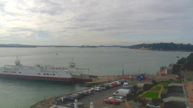 A coastal scene featuring a harbor with a large ferry docked at the pier. The ferry is mostly white with a red bottom and has markings on its side. In the foreground, there is a parking lot with various cars, and a small building nearby. The water is calm and reflects the overcast sky, which has soft clouds. In the background, there are hills and a wooded area along the shore, with a few boats visible on the water.