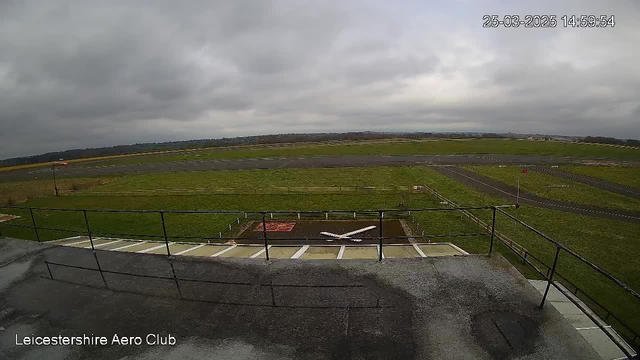 A wide view of an airfield under a cloudy sky. The foreground shows a low railing in front of a grassy area, with a large taxiway marked by white lines. In the center, a white aircraft is positioned on the ground next to a small red area. The background features an expanse of grass and a paved runway, leading to distant hills. The atmosphere appears overcast, indicating a dull weather condition.