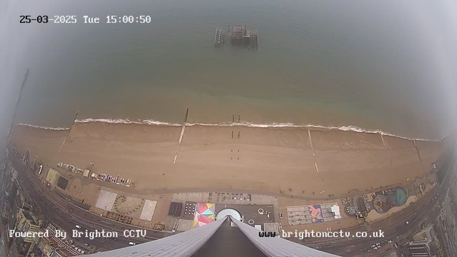 Aerial view looking down at a beach scene with light brown sand meeting the blue ocean. A pier extends into the water, with several small structures visible on the sand and in the ocean. There are people walking along the beach, and a promenade lined with buildings and vehicles is in the foreground. The date and time stamp indicates it is Tuesday, March 25, 2025, at 3:00 PM.