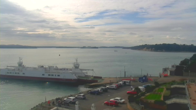 A view of a harbor with a ferry docked on the left. The ferry is white and red, labeled "GIVE WAY TO MAIN FERRY." The water is calm, reflecting the cloudy sky. In the foreground, there is a parking lot with several cars, predominantly white and red, and some people are seen near the dock. On the right, there is a landscaped area with grass and stairs. The background features distant hills and a few boats in the water.