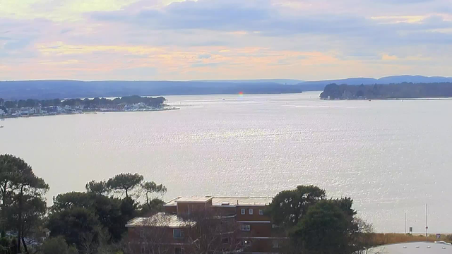 A scenic view of a calm body of water with a shimmering surface reflecting the sunlight. In the distance, there are small boats on the water, with a shoreline featuring houses and trees. The sky is partly cloudy with soft pastel colors, indicating a serene atmosphere. The scene captures the transition of light as the sun appears to be setting or rising on the horizon.