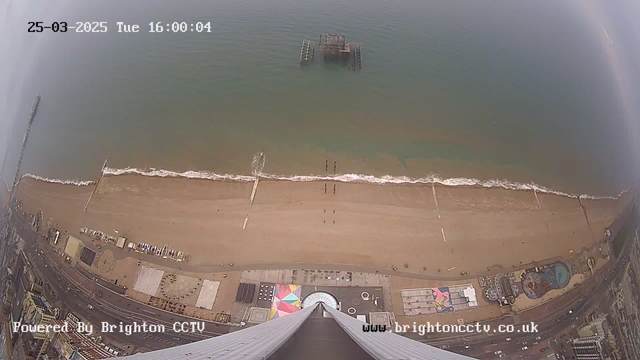 Aerial view of a sandy beach with gentle waves lapping at the shore. In the center, there is a partially submerged pier structure visible in the water. The beach is lined with colorful beach huts and a few people are walking along the sand. To the right, there is a swimming pool area next to some buildings. The sky is overcast, and the scene indicates a coastal environment.