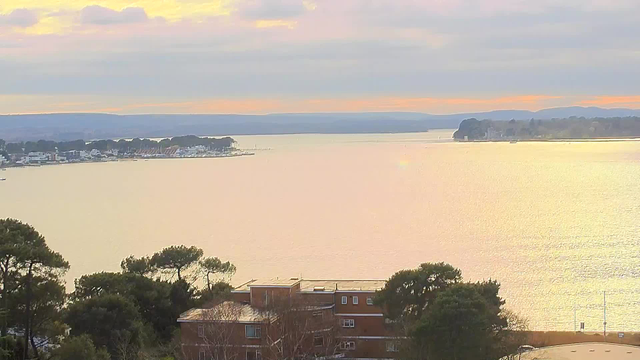A serene view of a lake at sunset, with soft light reflecting on the water's surface, creating a golden hue. In the foreground, a few trees are visible, along with a building made of bricks. The background features a distant shoreline lined with various structures and boats, while the horizon is framed by gentle hills under a cloudy sky. The overall atmosphere conveys calmness and tranquility.