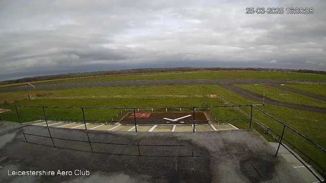 A wide view of an airstrip from the perspective of a tower. The foreground features a railing with a concrete surface. Below, there is a small helipad marked by a white cross on a brown field. The background shows a large, green grassy area with a cloudy sky above. There are some distant structures and a strip of asphalt to the right, with a partially blurred edge along the left side of the image.