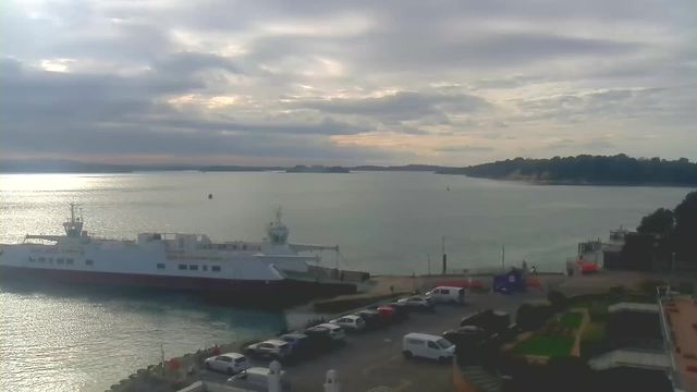 A serene view of a calm waterfront featuring a large white ferry docked at a quay. The water reflects soft hues of light, while the sky is filled with scattered clouds, creating a dreamy atmosphere. In the foreground, there are several parked vehicles along a roadway, and a green landscaped area can be seen to the right. The distant shoreline is lined with trees, and small boats can be spotted further out in the water.
