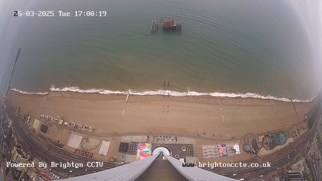 Aerial view of a beach with golden sand and gentle waves. In the foreground, there are colorful beach umbrellas and structures along the shore. A pier extends into the water, with a partially submerged structure visible nearby. The scene is under a cloudy sky, indicating an overcast day, with a few people walking along the beach. The timestamp at the top indicates the date and time.