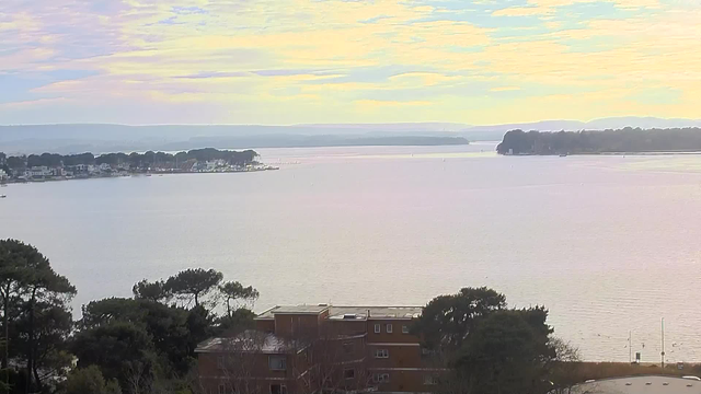 A serene view of a calm body of water extending towards the horizon, with light reflecting off the surface. In the distance, there are trees and hills, creating a gentle backdrop. On the left, a shoreline with houses can be seen, while a few boats are scattered across the water. The sky is filled with soft pastel colors, indicating an early morning or late afternoon setting. The foreground features some tree foliage and a building partially obscured by trees.