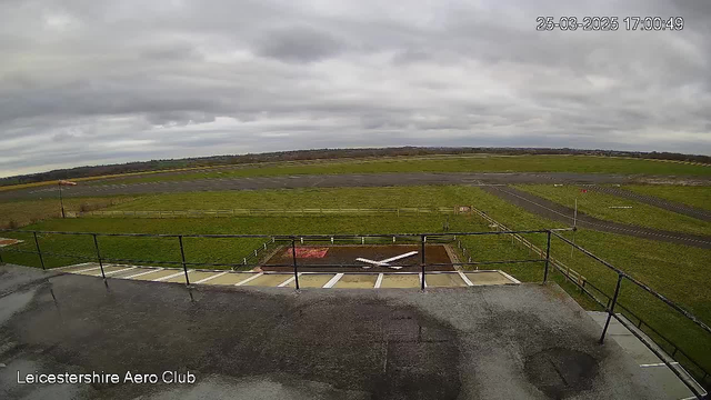 A cloudy sky appears above a wide-open grassy field. The view is from a high vantage point with a rail fence visible in the foreground. Below, there is a runway with a white X marking a designated area. In the distance, multiple runways can be seen along with a few scattered trees and farmland. The atmosphere is calm, suggesting a quiet day at the Leicestershire Aero Club.
