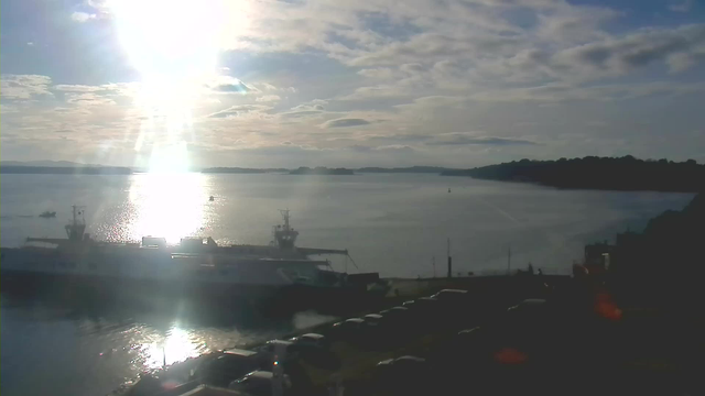 A peaceful waterfront scene with a ferry docked near the shore. The sun shines brightly in the sky, casting a reflective glare on the water's surface which also shows light ripples. In the background, there are rolling hills and several small boats. Several parked cars can be seen in the foreground along the waterfront. The sky is partly cloudy with a soft blue hue.