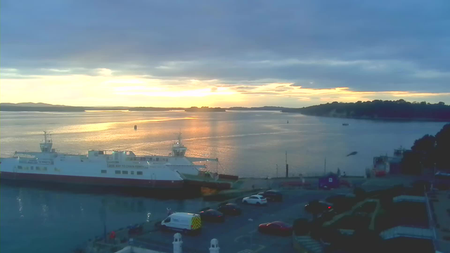A scenic view of a harbor at sunset. In the foreground, a large ferry boat is docked at the pier, with its side showing the words "GREAT WAY TO CHAIN FERRY". A few vehicles are parked nearby. The water reflects shades of orange and gold from the sunset, with gentle ripples moving across the surface. In the background, distant islands and hills are silhouetted against the sky, which is partly cloudy with varying shades of gray and blue.