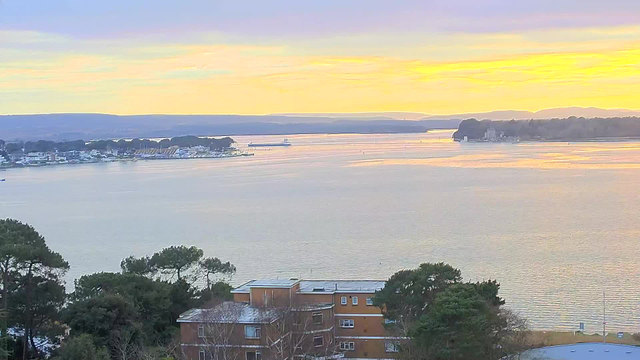 A scenic view of a water body at sunset, with a warm orange and yellow sky reflecting on the surface of the water. In the foreground, there are several trees and buildings, while the shoreline features docks with boats. The background includes distant hills and a calm waterway that leads further into the landscape. The overall atmosphere is tranquil and picturesque.