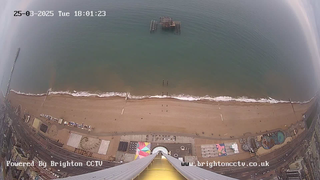 Aerial view of a beach and coastline. The sandy beach extends along the bottom of the frame, with gentle waves lapping at the shore. In the water, a partially submerged structure is visible, likely a pier. Colorful beach huts and a funfair area with various attractions can be seen on the left. The image is timestamped at 18:01 on March 25, 2025.
