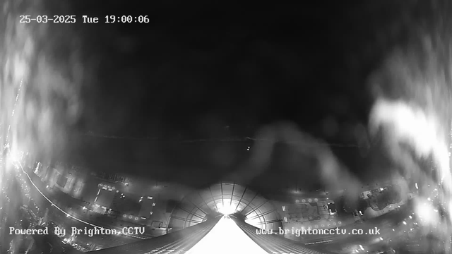 A dimly lit black and white image from a webcam shows an elevated view looking down a long, white path. On both sides of the path, the outlines of buildings and streetlights can be seen, partially obscured by foggy conditions. The top right corner displays the date and time: "25-03-2025 Tue 19:00:06." The bottom of the image includes text indicating the source: "Powered By Brighton CCTV" and a website link.