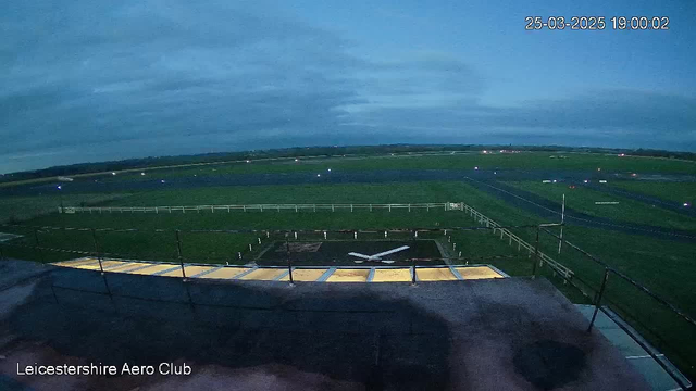A partially cloudy evening sky with a gradient from light to dark blue is visible above a green field. The foreground features a runway with a large white arrow symbol and a lighted pathway leading towards it. In the distance, lights can be seen along the runway and surrounding area, indicating activity at the Leicestershire Aero Club. The image has a timestamp showing March 25, 2025, at 19:00:02.