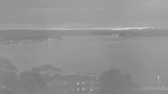 A grayscale image of a calm body of water reflecting overcast skies. In the distance, there are small lights from structures along the shoreline, indicating a settlement. A few trees are visible in the foreground, partially obscuring a building that is situated on the right side of the image. Overall, the scene conveys a tranquil and somewhat muted atmosphere.