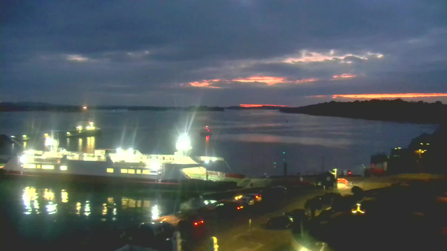 A dimly lit harbor scene at twilight. A large ferry boat is docked on the left, with bright lights reflecting off the calm water. The sky is mostly cloudy, with hints of orange and pink along the horizon as the sun sets. On the right side, there are parked cars near the shoreline, and faint lights from buildings are visible in the background.