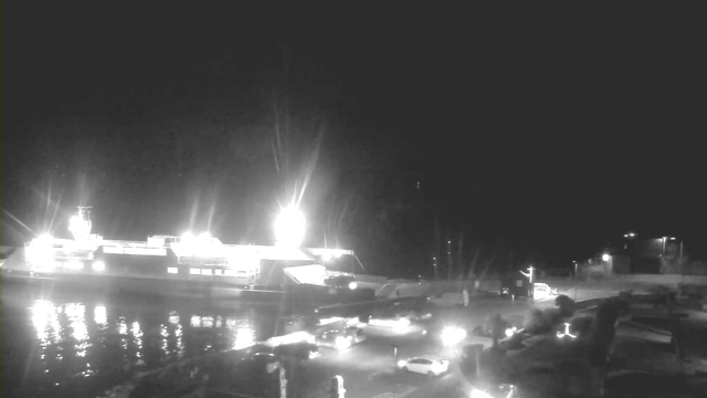 A dimly lit scene at a harbor during nighttime. A ferry is docked, illuminated by bright lights that create a reflection on the water's surface. Several vehicles are parked nearby, and some people can be seen walking along the waterfront. The atmosphere is calm, with minimal activity and a dark sky above.