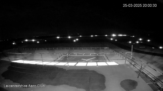 A dark scene showing a clear, open area at night with a few distant lights illuminating the surroundings. In the foreground, there is a rectangular area with a light reflecting off a surface, possibly indicating a runway or helipad, marked with an 'X.' A railing runs along the perimeter of this area. The background is mostly dark with faint lights seen at a distance. The image timestamp is visible in the top right corner, indicating the date and time.