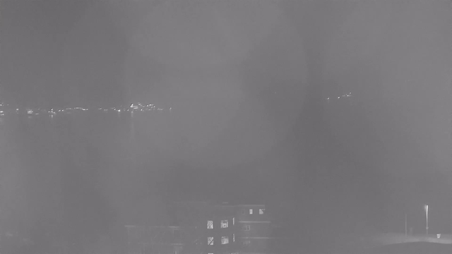 A dimly lit scene shows a body of water with a distant shoreline illuminated by scattered lights. In the foreground, there is a view of a building with some windows glowing faintly, while the overall atmosphere appears hazy and obscured, possibly due to fog or mist.