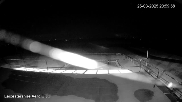 A dark scene captured by a webcam at the Leicestershire Aero Club. In the foreground, there are outlines of railings and flat surfaces, partially illuminated by a faint light. The background is mostly dark, with some distant lights suggesting a landscape beyond. The date and time displayed in the upper right corner indicate it is evening on March 25, 2025.