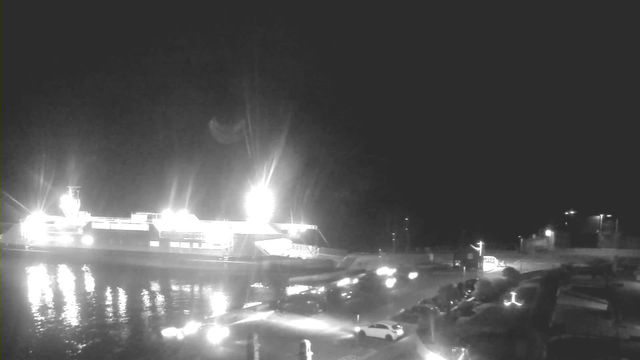 A brightly lit ferry docked at a harbor during nighttime. The water reflects the ferry's lights, creating shimmering patterns. In the foreground, several parked cars are visible along the waterfront, and faint lights from buildings and streetlights are seen in the background, contributing to the nighttime scene.