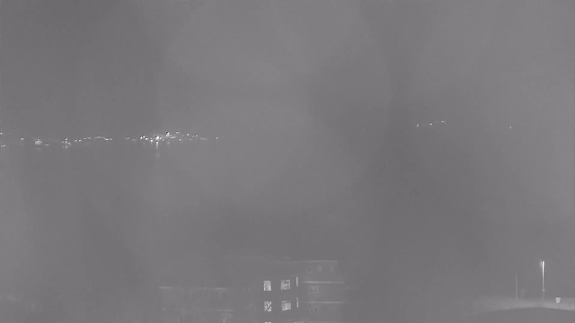 A low-contrast black and white image depicting a body of water with faint lights reflecting on its surface in the distance, suggesting the presence of a shoreline or structures. In the foreground, there are outlines of buildings, with a few windows illuminated, providing some contrast against an overall blurred and hazy background.