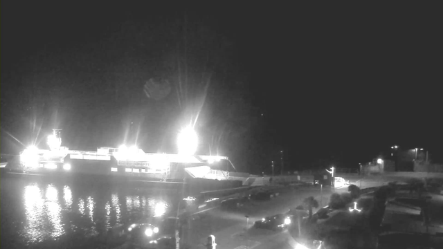 A dimly lit scene of a harbor at night, featuring a large illuminated ferry docked at the pier. Surrounding the ferry, the water reflects the bright lights. On the dock, there are several parked vehicles and faint streetlights illuminating the area. The background is mostly dark with some distant structures outlined against the night sky.