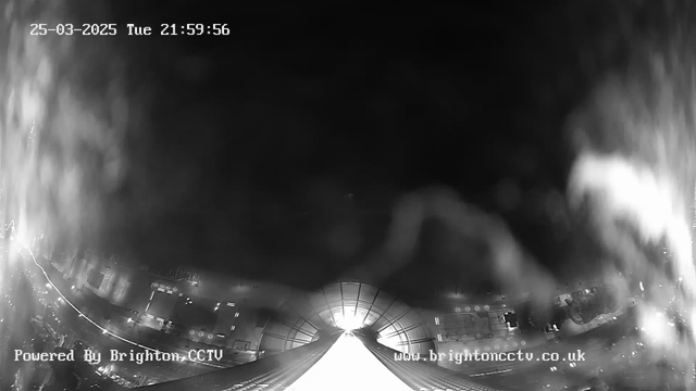 A black and white webcam image showing a nighttime view. The lower part displays a brightly lit area resembling a long, sloped surface leading into darkness. There are faint outlines of buildings and a road with lights visible in the background. The date and time are displayed at the top of the image, with the words "Powered By Brighton CCTV" in the lower left corner. The overall scene conveys a sense of depth, with the light suggesting an entrance or tunnel leading away from the viewer.