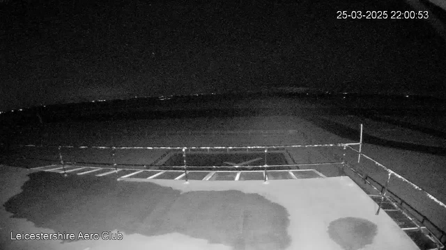A dark scene showing a view from a webcam at the Leicestershire Aero Club. In the foreground, there is a flat surface with visible markings, possibly a runway or landing area. A rail or barrier is present along one side, and faint lights are visible in the distance, suggesting a horizon or distant buildings. The overall ambiance is dim, indicative of nighttime conditions. The timestamp indicates it is March 25, 2025, at 10:00 PM.