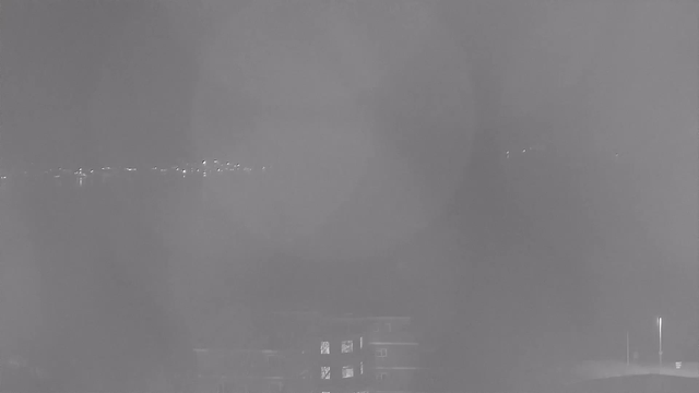 A black-and-white image shows a view of a faint cityscape at night, characterized by blurred outlines of buildings and distant lights. The scene is hazy, suggesting fog or mist, with shapes and patterns indistinct due to low visibility. A structure with several lit windows is visible in the foreground, and a line of soft lights can be seen in the background, likely from a roadway or distant buildings.