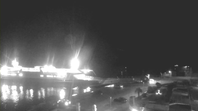 A illuminated ferry is docked at a harbor at night, with bright lights reflecting on the water. There are a few parked cars visible in the foreground, and additional lighting from the shore and parking area suggests a busy space. The background is dark, highlighting the lights from the ferry and surrounding structures.
