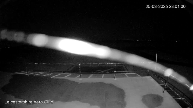 A dimly lit scene shows an outdoor area at night, with a flat surface that appears to be wet. There is a blurred streak across the image, possibly from water on the camera lens. In the background, there are faint outlines of a fence or railing. The scene is mostly dark, with some distant lights potentially visible far in the background. The lower left corner displays the text "Leicestershire Aero Club" in white. The date and time in the upper right corner read "25-03-2025 23:01:00."