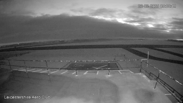 A black and white webcam image showing a cloudy sky at dawn over a flat landscape. A fenced area, likely a helipad, is visible in the foreground, marked with white lines. In the background, there are distant fields and low hills, partially obscured by clouds. The scene appears calm and still, with no vehicles or people present.