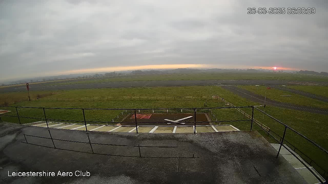 A cloudy sky is visible above an airfield at dawn. In the foreground, there is a metal railing surrounding a viewing platform. Below, a grassy area features a marked landing area with a large white X and an adjacent red patch. To the right, there is a section of the airfield with taxiways and a small red sign. The sun is rising on the horizon, creating a soft glow in the sky.