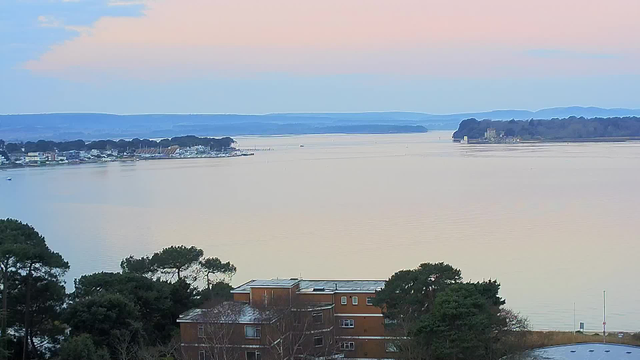 A tranquil view of a body of water during twilight, with a gradient sky transitioning from soft pink to light blue. In the foreground, there are trees and a multi-story brown building. Across the water, a small marina with boats is visible, along with distant hills and islands on the horizon. The surface of the water appears calm and reflective, enhancing the serene atmosphere of the scene.