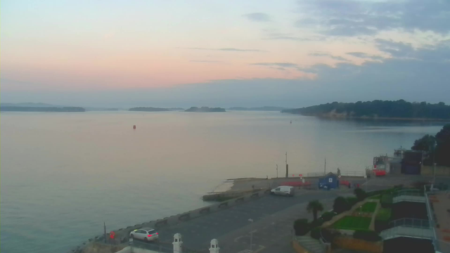 A tranquil waterfront scene during dawn, featuring calm water reflecting soft pastel colors in the sky. In the foreground, there is a parking area with a few vehicles, and a pathway leads towards the water. On the left, a small dock can be seen, with a couple of vessels. In the distance, there are small islands and a shoreline covered in greenery. The atmosphere is peaceful and serene, with gentle clouds scattered across the sky.