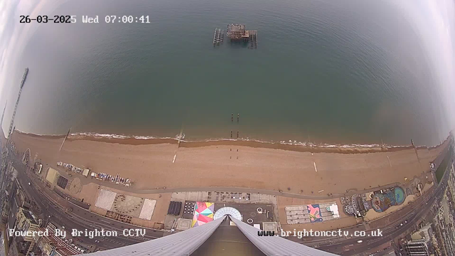 A high-angle view of a beach with a sandy shore and calm, blue water. In the distance, a pier extends out over the water. There are several structures visible on the beach, including a colorful circular design and some boats. The sky is mostly cloudy but bright. The scene captures a quiet early morning atmosphere.