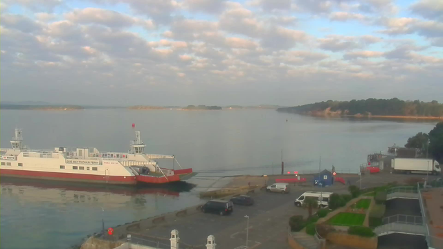 A view of a calm water body with a cloudy sky above. In the foreground, a ferry with a white upper deck and red lower hull is docked at a pier, with a sign that reads "GIVE WAY TO CHAIN FERRY." Several vehicles, including a white truck and a dark-colored car, are parked nearby. The shore features patches of green grass and a few trees, while a distant landmass is visible across the water. The serene atmosphere is complemented by reflections on the water's surface.